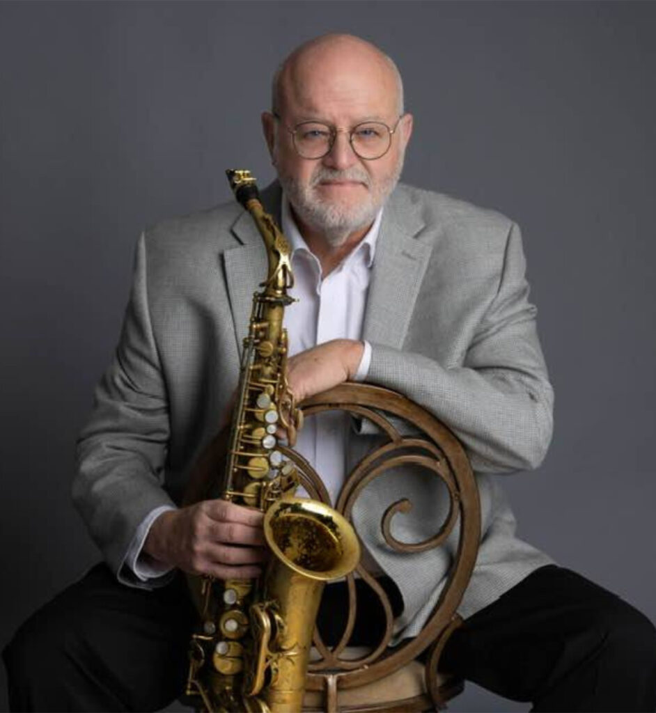 Steve Williams sitting backward in a chair dressed in a grey blazer and holding his saxophone