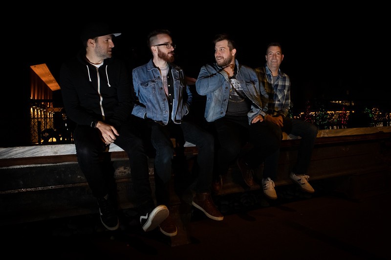 keeton band members sitting on rooftop wall at night with city in background.